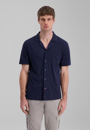 Man wearing a navy short-sleeve button-up shirt and light gray cargo shorts, standing against a plain gray background.