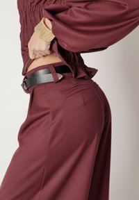 Burgundowe spodnie z gładkiego materiału, wysoki stan i czarny skórzany pasek z srebrną klamrą i akcentami.