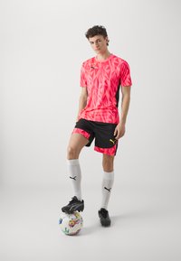 Maglia sportiva a maniche corte con motivo rosa brillante e pantaloni neri con dettagli rosa, abbinati a calze bianche e scarpini neri, in piedi su un pallone da calcio multicolore.