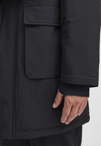 Chaqueta exterior negra con una textura suave, que presenta un gran bolsillo y un puño en las mangas. La tela parece resistente al agua y duradera.
