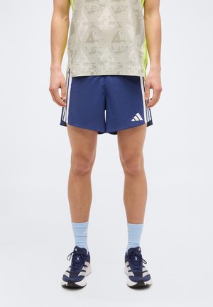 Άνδρας με μπλε αθλητικό σορτς Adidas με λευκές ρίγες, ανοιχτό μπλε κάλτσες και σκούρα μπλε παπούτσια τρεξίματος, στέκεται μπροστά σε λευκό φόντο.