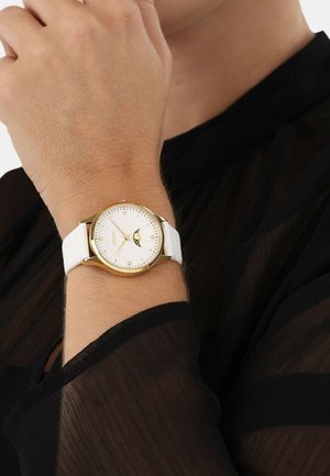 Goldfarbene Armbanduhr mit weißem Lederarmband, rundem Ziffernblatt mit goldfarbenen Akzenten und einer Mondphasenanzeige. Schwarzes, transparentes Oberteil teilweise sichtbar.