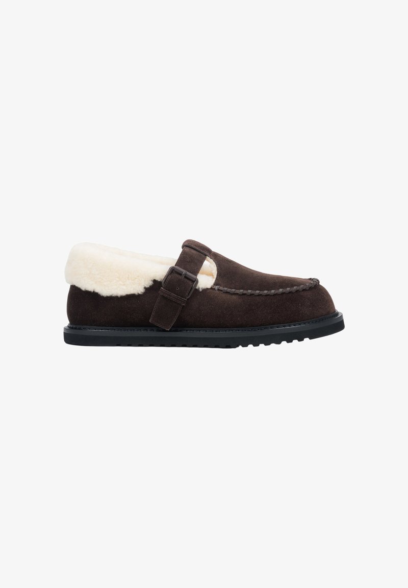 Bruin suède slipper met een witte nepbont lining, voorzien van een gespband en een gestructureerde zwarte rubberen zool. Klassiek moccasin ontwerp.