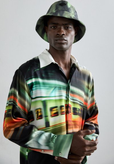 Langærmet skjorte med et farverigt abstrakt mønster i grøn, orange og sort; har en hvid krave og knapmanchetter. Kombineret med en grøn bucket hat.