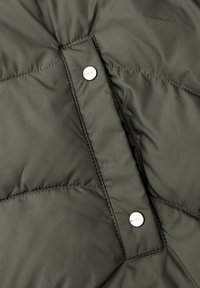 Tejido de chaqueta acolchada en verde oscuro con textura acolchada. Cuenta con cierres de presión plateados y un detalle de costura visible.