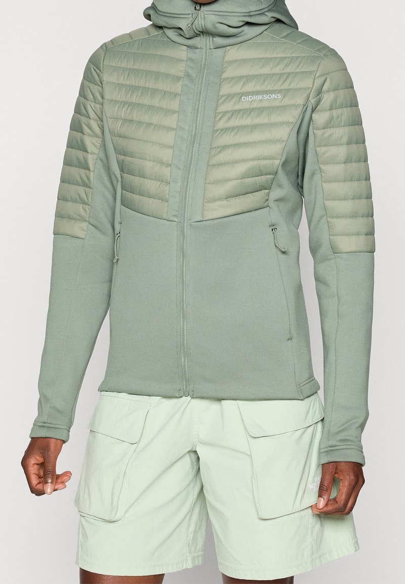 Personne portant une veste matelassée vert clair avec fermeture éclair devant et capuche, associée à un short cargo clair avec plusieurs poches.