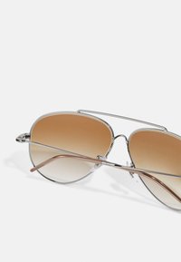 Óculos de sol estilo aviador com lentes castanhas em degradê, armação de metal prateado, almofadas nasais transparentes e detalhe castanho na têmpora.