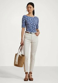 Γυναίκα με μπλε φλοράλ μπλούζα, λευκά παντελόνια, καφέ σανδάλια, που κρατάει μια υφαντή τσάντα με λευκό ύφασμα μέσα.