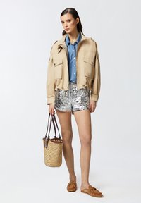 Giacca beige cropped con due tasche frontali, pantaloni corti metallici argento, sandali marroni slip-on e una borsa di paglia a disegni.