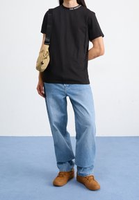 Černé tričko s krátkým rukávem a bílým textem na límci, volné modré džíny a tanové boty. Také obsahuje tanovou crossbody tašku.