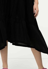 Schwarzes schichtetes Kleid mit fließender Silhouette, aus leichtem Stoff gefertigt. Es endet auf halber Wade mit einem Saum, der nach oben verläuft.
