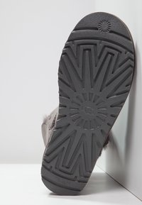 Suola grigia di calzature con superficie in gomma testurizzata che presenta un motivo a raggi di sole, bordi curvi e logo "UGG" impresso al centro.
