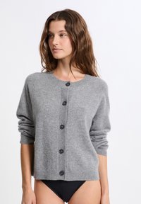 Femme avec de longs cheveux ondulés portant un cardigan gris clair à boutons et une culotte noire, tournée vers la gauche contre un fond blanc uni.
