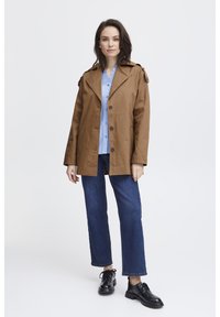 Brauner Baumwoll-Trenchcoat mit Schulterriegeln, Knopfleiste, über einem hellblauen Hemd getragen, kombiniert mit blauen Jeans und schwarzen Schuhen.