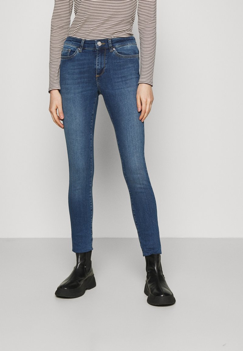 Jeans ajustados de mezclilla azul oscuro con una altura media, que cuentan con bolsillos frontales y un cierre de botón. Combinados con botas negras hasta el tobillo.