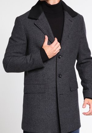 Hombre vistiendo un abrigo de lana gris oscuro con cuello negro y un cuello alto negro, ajustando el delantero del abrigo con una mano.
