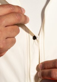 Veste blanche légère avec fermeture éclair et détail de bouton, dotée d'une doublure en mesh respirant visible près du col.