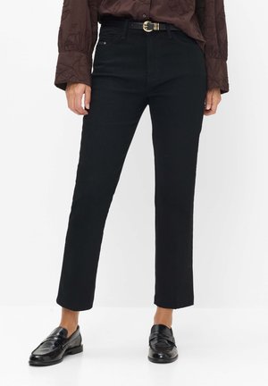 Kvinde iført tætsiddende sorte bukser med sort bælte og sorte læder loafers, kombineret med en mørkebrun bluse med lange ærmer og teksturerede detaljer.