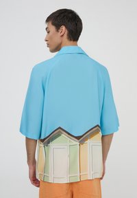 Camicia a maniche corte in tessuto azzurro chiaro, con una stampa colorata di un bungalow sulla spiaggia all'orlo, colletto strutturato e texture liscia.