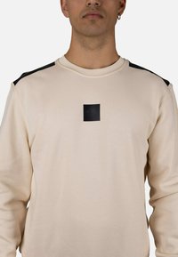 Sudadera beige con detalles negros en los hombros. Presenta un parche cuadrado con el logo negro en el pecho. Hecha de tela suave y texturizada.