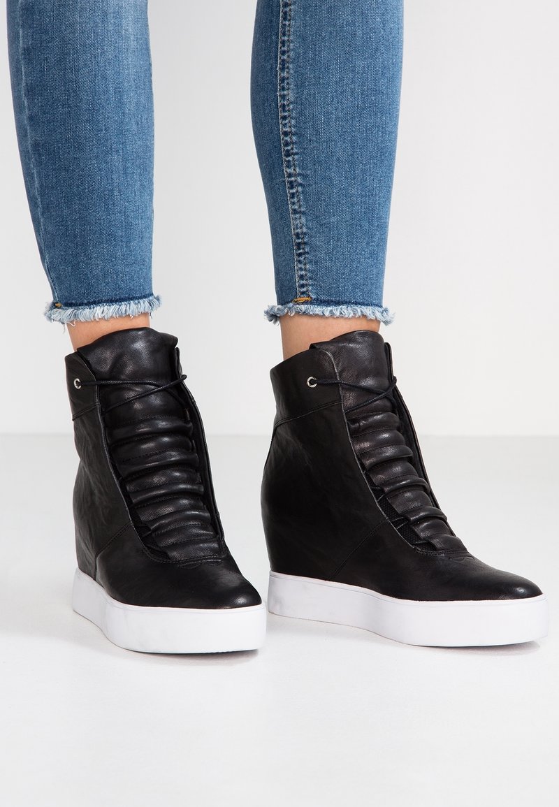 Černé klínové tenisky s plisovaným koženým svrškem, bílou gumovou podešví a šněrováním vpředu. Stylizované s roztrhanými kotníkovými džíny.