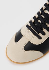 Scarpa sportiva nera e beige con una tomaia in pelle liscia, suola in gomma e lacci neri. Dettagli di cucitura notevoli lungo il design.