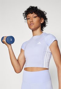 Svetlofialový športový crop top s krátkymi rukávmi, ozdobený tromi bielymi pruhmi po boku, v kombinácii s vysoce pásovými legínami a modrou činkou.