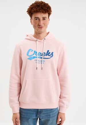 Jonge man met krullend rood haar en sproeten, die een lichtroze hoodie met blauwe tekst "Creeks Original Since 1974" en een blauwe jeans draagt.