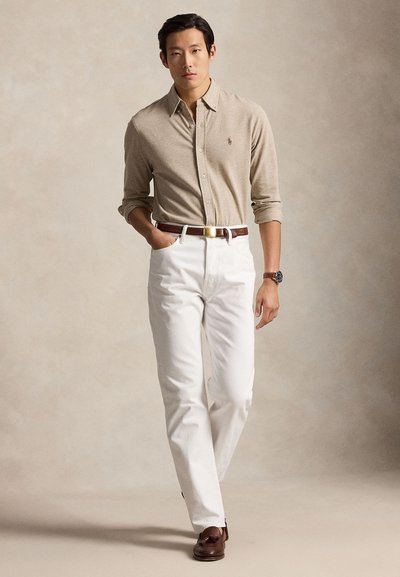 Moški, ki nosi bež srajco z gumbi, bele hlače, rjavi pas, uro in rjave loaferje, stoji z roko v žepu.