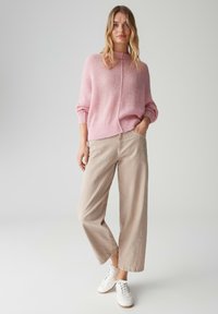 Maglione rosa chiaro lavorato a maglia con collo alto, abbinato a pantaloni beige a gamba larga e sneakers bianche. L'outfit presenta una vestibilità morbida e una texture soffice.