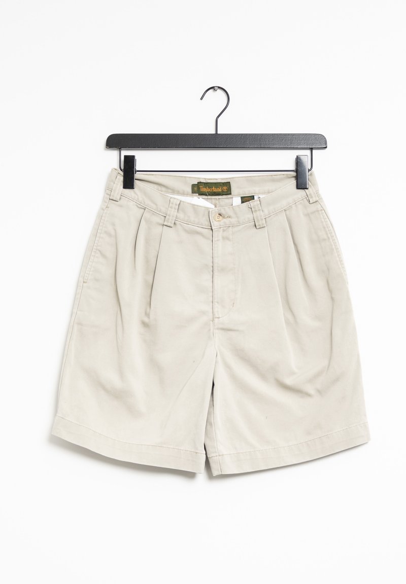 Shorts en coton beige avec plis, passants pour ceinture et fermeture à bouton sur le devant. Suspendus à un crochet noir sur un fond blanc.