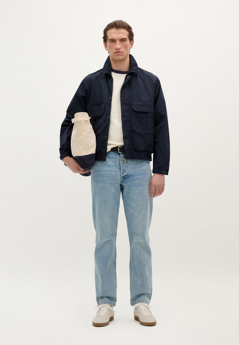 Veste navy avec un col de chemise, deux poches avant, t-shirt crème, jean bleu clair et baskets beiges avec une semelle blanche. Sac beige tenu sous le bras.