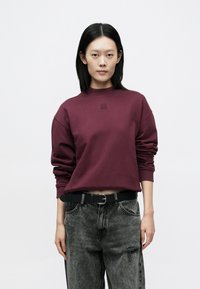 Vinrød genser med ribbet rund hals og senkede skuldre, kombinert med slitte svarte jeans og et svart belte. Har en diskret logo.