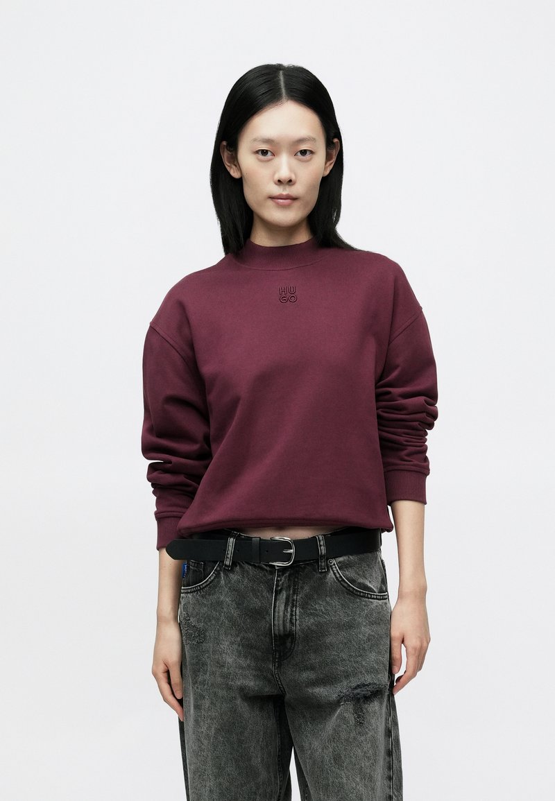 Vinrød genser med ribbet rund hals og senkede skuldre, kombinert med slitte svarte jeans og et svart belte. Har en diskret logo.