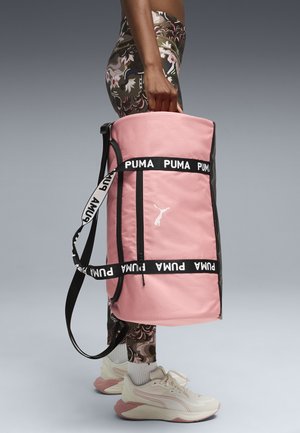 Persona che indossa leggings floreali e sneaker bianche, portando di lato una grande borsa da viaggio rosa Puma con cinghie nere con il logo.