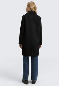 Cappotto nero con finitura testurizzata, maniche lunghe e ampia colletto. Presenta una singola apertura centrale sul retro, abbinato a jeans blu svasati.