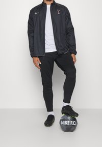 Nike vindjacka i svart med Tottenham-logotyp, i kombination med svarta åtsittande byxor och svarta fotbollsskor, stående på en svart Nike-fotboll.