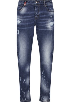 Mees, kes kannab tuhmunud siniseid kulunud teksapükse ja valgeid, beeže ning musti vabaajajalatsid, seisab külje pealt.