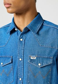 Camicia in jeans blu con colletto a punta, bottoni a pressione e due tasche sul petto. Presenta cuciture gialle a contrasto e un logo a patch.