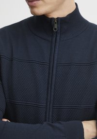 Tamsiai mėlynas zip-up megztinis su aukštu kalaru, turintis nertą tekstūrą ir įspaustą deimantų raštą per krūtinę. Užtrauktukas priekyje.