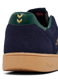 Marinblå mockaskor med gröna läderdetaljer på hälen. Har en gyllene varumärkeslogotyp och chevrondetaljer, samt en strukturerad gummisula.