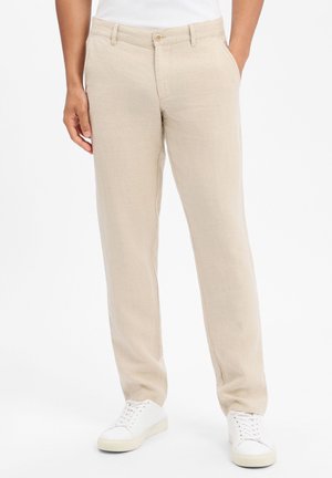 Beige broek met rechte pijpen, gedragen met witte sneakers en een wit shirt in de broek gestopt, handen rustend langs het lichaam en in de zak.