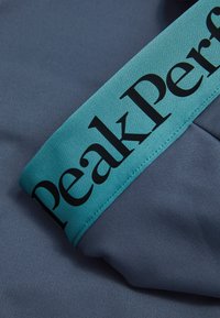 Blåt stof tæt på med en teal elastisk talje og "Peak Performance" i sort tekst. Tekstureret overflade med glat finish.