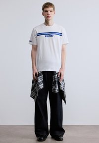 Bela bombažna majica z modrimi črtami in logotipom, kombinirana s črnimi širokimi jeans hlačami in črno-belo karo srajco, zvezano okoli pasu.