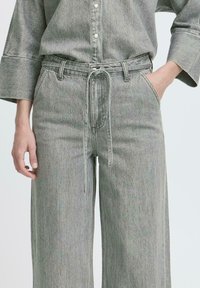 Szürke farmer anyagú, bőszárú nadrág húzózsinóros derékrésszel, sima textúrával és kontrasztos varrással. Látható első zsebek és övhurkok.