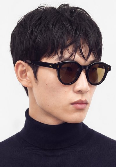 Jeune homme aux cheveux noirs courts, portant des lunettes de soleil rondes et foncées et un pull à col roulé noir, regardant sur le côté.
