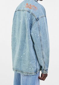 Ljusblå denimjacka med stänkmönster och orange text på ryggen. Har en avslappnad passform, långa ärmar och knäppning vid ärmsluten.