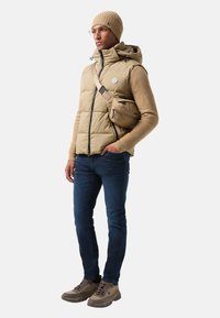 Beige polstret vest med hætte over en tan sweater, mørkeblå jeans og brune vandrestøvler. Tilbehør inkluderer en brun crossbody taske og en strikket hat.