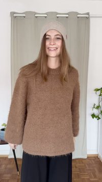 Brązowy, miękki sweter z długimi rękawami i zaokrąglonym dekoltem, zestawiony z jasnobeżową, dzierganą czapką. Prosty, luźny fason.