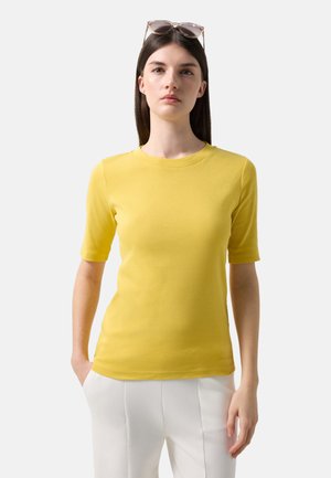 Frau mit glatten braunen Haaren trägt ein gelbes kurzärmeliges Oberteil, weiße Hose und eine Sonnenbrille, die auf ihrem Kopf sitzt, und steht vor einem einfarbigen Hintergrund.